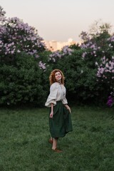Young pretty caucasian woman in romantic outfit walking in blooming spring garden. Artistic outdoor photo of beautiful red-haired girl posing in fragrant lilac blossom.