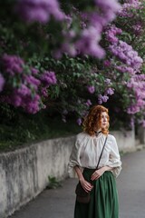 Young pretty caucasian woman in romantic outfit walking in blooming spring garden. Artistic outdoor photo of beautiful red-haired girl posing in fragrant lilac blossom.