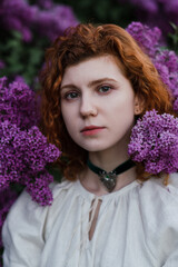 Young pretty caucasian woman in romantic outfit walking in blooming spring garden. Artistic outdoor photo of beautiful red-haired girl posing in fragrant lilac blossom.