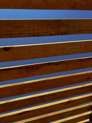 Modern wooden pergola roof with evenly spaced slats against a clear blue sky. The architectural structure creates a dynamic geometric pattern with light and shadow, highlighting natural wood texture