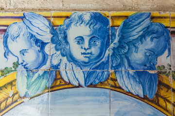 a close-up of a vibrant, historical azulejo panel featuring three cherubic faces in blue and white, positioned above a decorative golden arc and lush green foliage