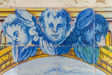 a close-up of a vibrant, historical azulejo panel featuring three cherubic faces in blue and white, positioned above a decorative golden arc and lush green foliage