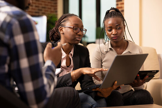 African American startup team work together in reviewing documents and digital report on laptop during financial project discussion. Professional woman explains marketing strategy plans to colleagues.