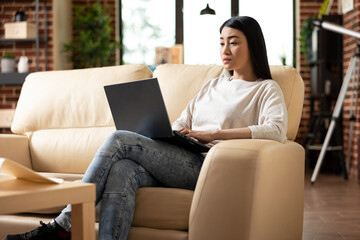 Professional female adult sitting on couch with laptop on lap, completing company reports from...