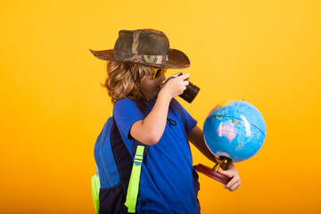 Cute blond kid wearing explorer hat and backpack on studio. Child explorer and adventure concept. Kid tourists hold world globe isolated on yellow background.