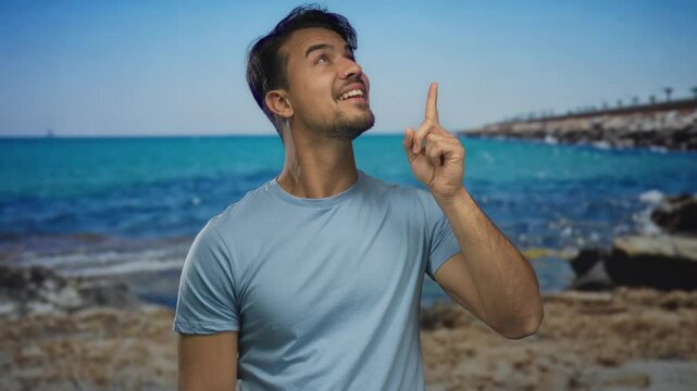 Young hispanic man smiling at a seaside beach, pointing confidently with the ocean and blue sky in the background, conveying a sense of joy and leisure outdoors.
