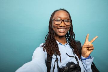 African american female vlogger giving peace sign to her online audience, recording herself in studio. Pov of excited black woman photographer making two finger, v gesture towards the camera.