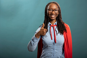 Cheerful black heroine giving a thumbs up, portraying positivity and agreement. African american woman in red cape showing approval gesture and looking at camera with bright smile.