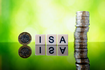 ISA tax-free savings concept with letter tiles, single pound coin and tall stack of sterling coins reflected on glossy surface against green bokeh background.