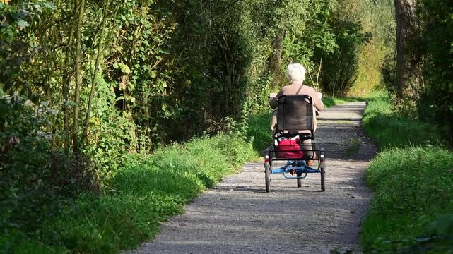 An elderly woman with grey hair rides a tricycle with a luggage rack on a dirt road among the trees. 