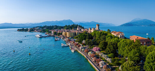 Fototapeta premium Aerial view of Sirmione, Italy, with boats, historic Scaliger Castle, waterfront promenade, and Lake Garda surrounded by mountains