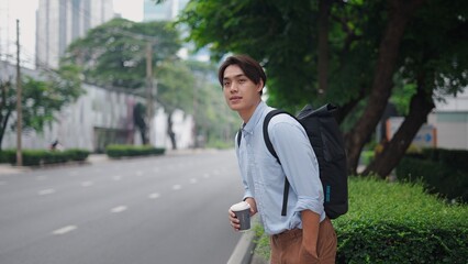 Young asian businessman waiting by roadside, holding coffee, carrying backpack