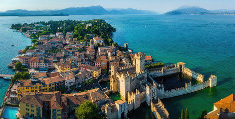 Aerial view of Sirmione, Italy, with boats, historic Scaliger Castle, waterfront promenade, and Lake Garda surrounded by mountains