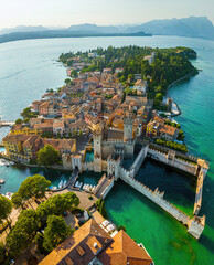 Aerial view of Sirmione, Italy, with boats, historic Scaliger Castle, waterfront promenade, and Lake Garda surrounded by mountains