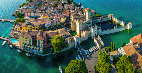 Aerial view of Sirmione, Italy, with boats, historic Scaliger Castle, waterfront promenade, and Lake Garda surrounded by mountains