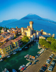 Aerial view of Sirmione, Italy, with boats, historic Scaliger Castle, waterfront promenade, and Lake Garda surrounded by mountains