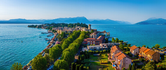 Aerial view of Sirmione, Italy, with boats, historic Scaliger Castle, waterfront promenade, and Lake Garda surrounded by mountains