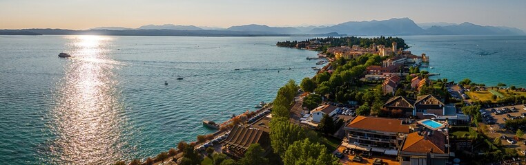 Aerial view of Sirmione, Italy, with boats, historic Scaliger Castle, waterfront promenade, and Lake Garda surrounded by mountains