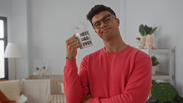 Young man in bedroom holding mug with text best dad ever, wearing glasses, red sweater, indoor setting, smiling, enjoying moment, suggesting family, fatherhood, interior decor.