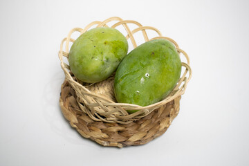 Two fresh green mangoes placed in a woven bamboo basket on a braided mat, set against a clean white background.