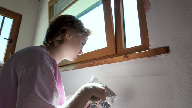 Teenage girl spreading putty across wall surface. Filling cracks with spackle, preparing walls for painting. Helping parents with home renovation. Slow motion sunny bright indoor footage.