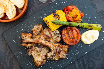 Closeup of appetizing lamb chops on ribs garnished with grilled corn, tomato slice, sweet pepper and asparagus on black slate serving board. Traditional mediterranean cuisine