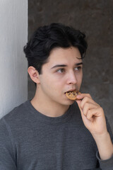 Apuesto joven sobre una pared gris comiendo galleta con chispas de chocolate con una expresión...