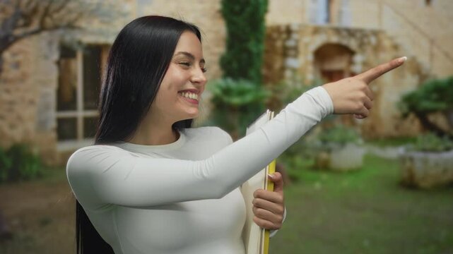 Woman smiling outdoors pointing and holding books near a rustic building with stone walls and lush greenery in the background capturing a cheerful academic scene.