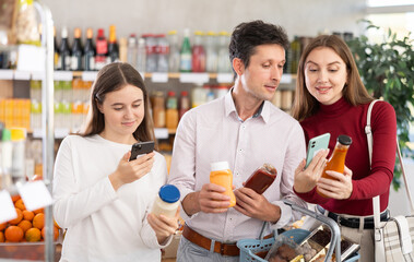 While shopping, family of customers doubts choice of sauce and studies information about composition of product in online application. Parents and daughter scan barcode on product label.