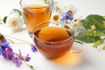 Aromatic herbal tea in glass cups, different flowers and green leaves on white wooden table, closeup