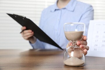 Deadline. Woman with hourglass and clipboard at wooden table in office, closeup
