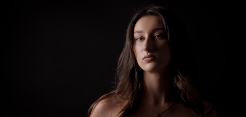 Fototapeta premium Studio portrait of a young Caucasian woman with long brown hair, looking serious with bare shoulders against a dark background