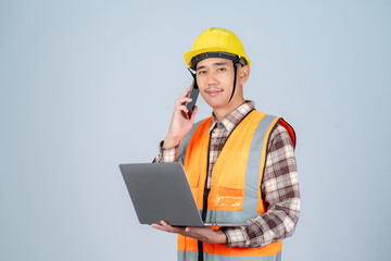 Smiling young Asian engineer or worker wearing yellow hard hat and reflective vest using laptop and smartphone on grey background.