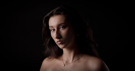 Naklejka premium Studio portrait of a young woman with long brown hair and bare shoulders, wearing a necklace, looking with a serious and intense expression against a dark background