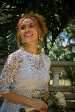 "Beautiful Latin bride with curly hair sitting on a park bench, holding a bouquet of fresh flowers and smiling joyfully on her wedding day. Dressed in a flowing white gown, she radiates happiness and 