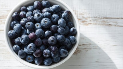 overhead view of washed and clean Blueberries in a white ceramic bowl sitting on a solid wood kitchen table with white sunlight coming in through a window on it 