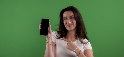 Young woman holding and pointing at a smartphone with blank screen on green background, perfect for...
