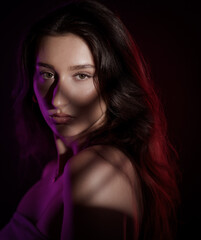 Studio portrait of a young woman with long brown hair, illuminated by purple and red lights casting...