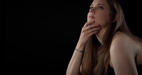 Thoughtful young woman is holding her chin and looking up on a dark background