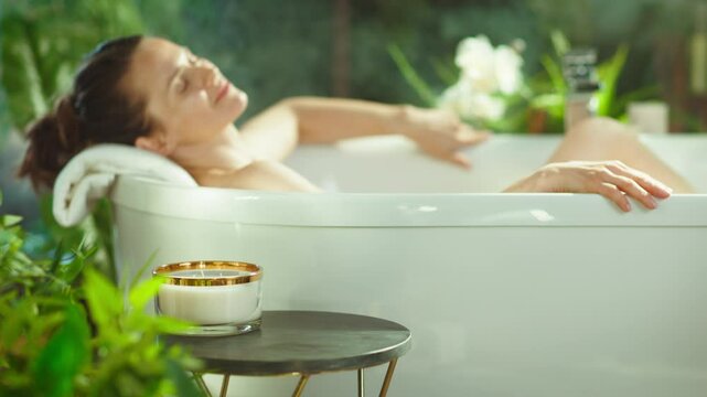 A blurred woman with hair in a bun relaxes in a white bathtub with a pillow, surrounded by soft light, green plants, & flowers. A candle on a table enhances this calm, luxurious home spa setting.