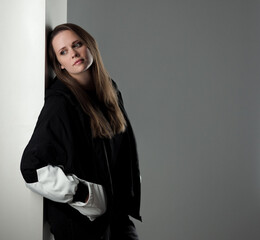 Young woman with long brown hair wearing black and white jacket is leaning against white wall and...