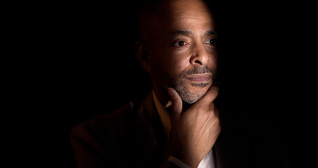Portrait of a thoughtful businessman with hand on chin, illuminated by a ray of light in a dark...