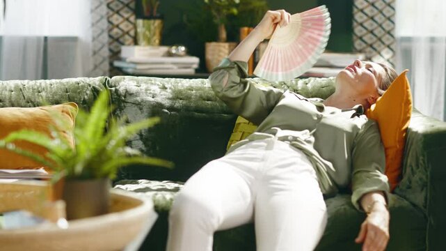A middle-aged woman on a green velvet sofa, fanning with a hand fan, eyes closed, seeking relief from heat in a sunlit living room. She may be experiencing exhaustion or hot flashes.
