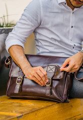 Fototapeta premium A man in a white shirt sits and opens a large brown leather shoulder bag, perfect for a leather products catalog design.