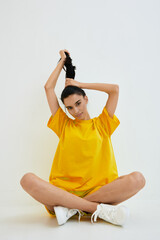 young woman in yellow oversized shirt sitting crossed legged on white floor tying her hair with a...