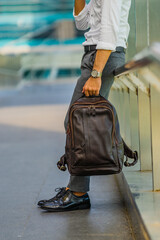 Fototapeta premium A man in a white shirt leans against an iron fence carrying a brown leather backpack, perfect for a leather products catalog design.