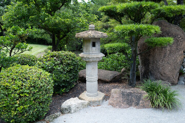 A traditional stone lantern stands among neatly trimmed bushes, rocks, and sculpted trees in a...