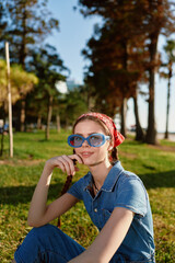 young woman wearing blue sunglasses and red headscarf sitting on grass in sunny park surrounded by...