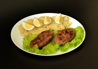 A plate of delicious lunch, mashed potatoes with sliced ​​chicken egg and two fried meat cutlets with lettuce leaf on a black background.