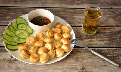 Large plate with fried dumplings, fresh sliced ​​cucumber and spicy sauce on a wooden table, a cup of tea and a fork nearby.